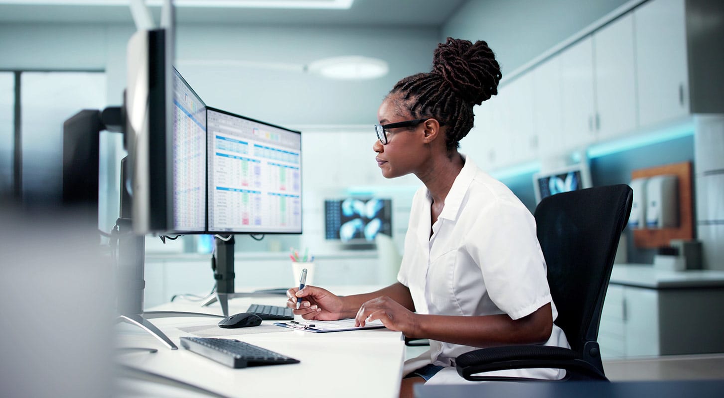 Female doctor in glasses and a white coat working at a desk, reviewing patient scheduling or data on two computer monitors.